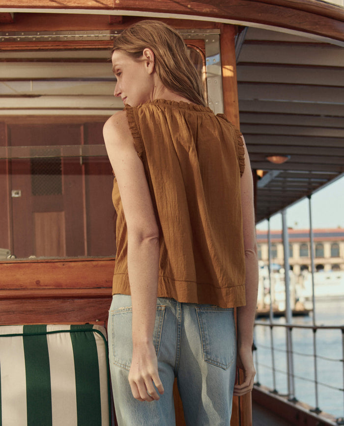 A woman wearing THE GREAT. SU26 The Vignette Top in Clay and light blue jeans stands on a boat deck, facing away from the camera.