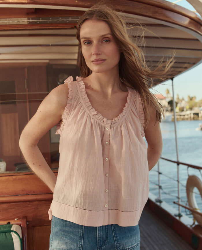 A woman wearing THE GREAT. SU26 The Vignette Top in Petal and denim jeans stands on a wooden boat, with water and buildings in the background.