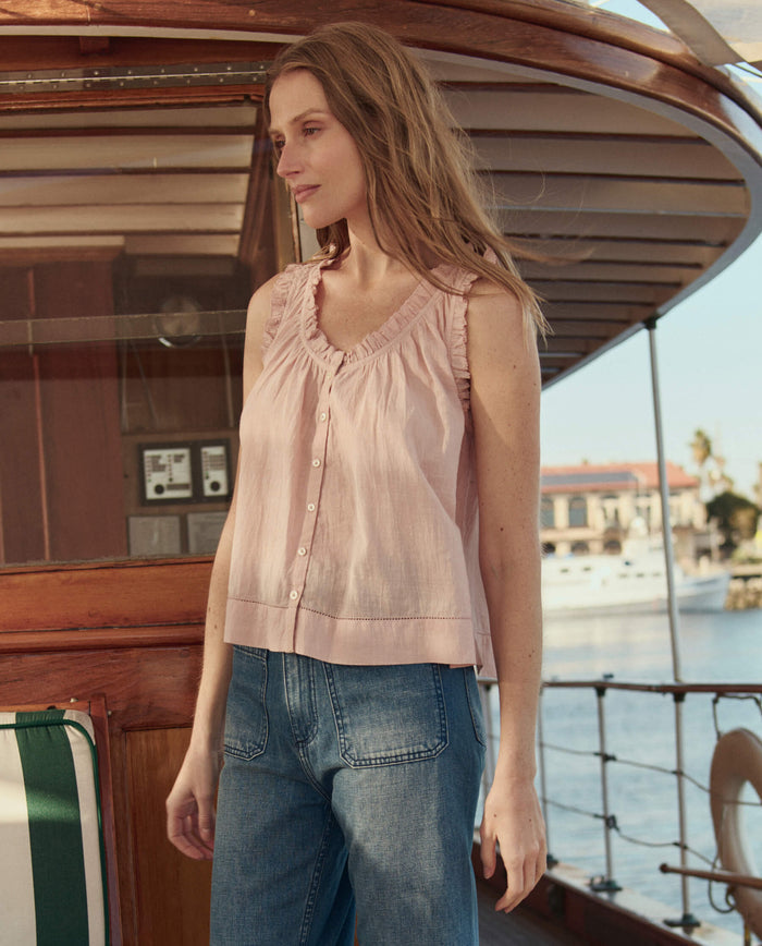 A woman with long hair stands on a wooden boat, wearing THE GREAT. SU26 The Vignette Top in Petal and blue jeans, with buildings and water in the background.