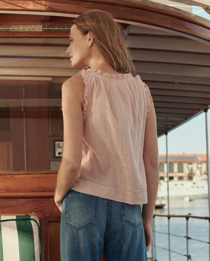A person with long hair stands on a boat facing away, wearing THE GREAT. SU26 The Vignette Top in Petal and blue jeans, with water and buildings in the background.