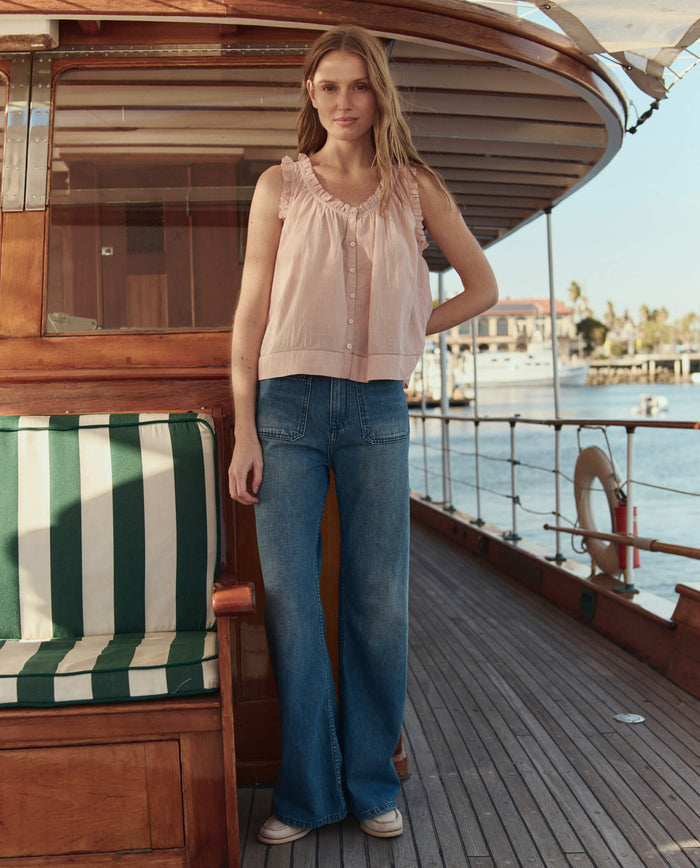 A woman in THE GREAT. SU26 The Vignette Top – Petal, paired with blue wide-leg jeans, stands on a wooden boat deck with water and city buildings in the background.