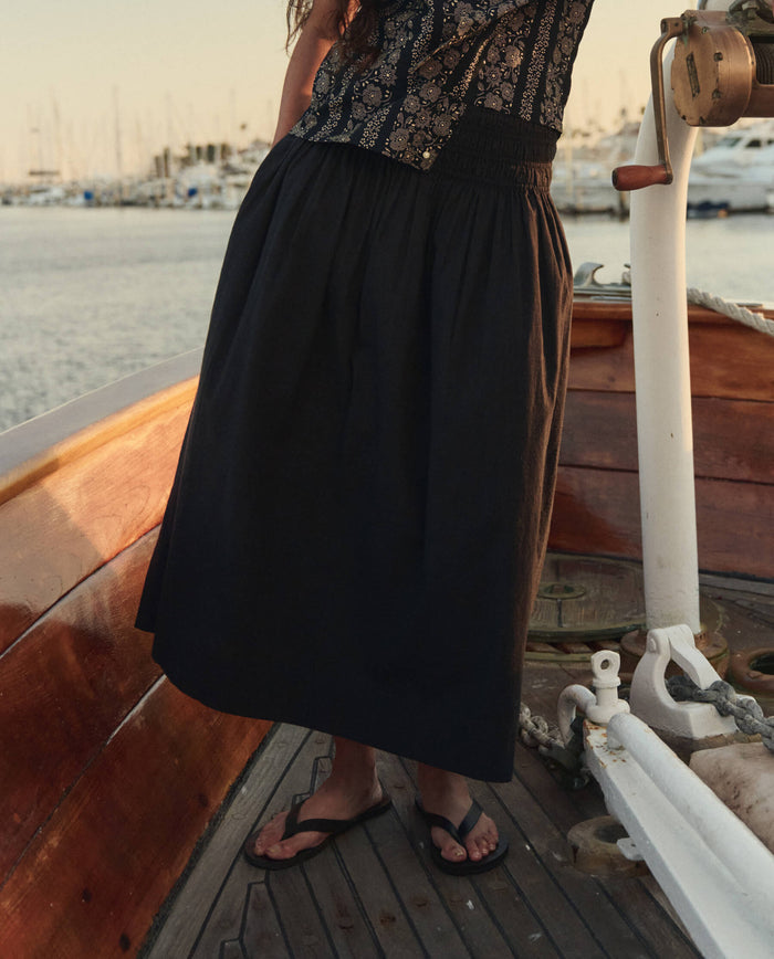 Wearing THE GREAT. SU26's The Viola Skirt in black, a person pairs it with a patterned top and flip-flops while standing on a wooden boat, marina and boats visible in the background.