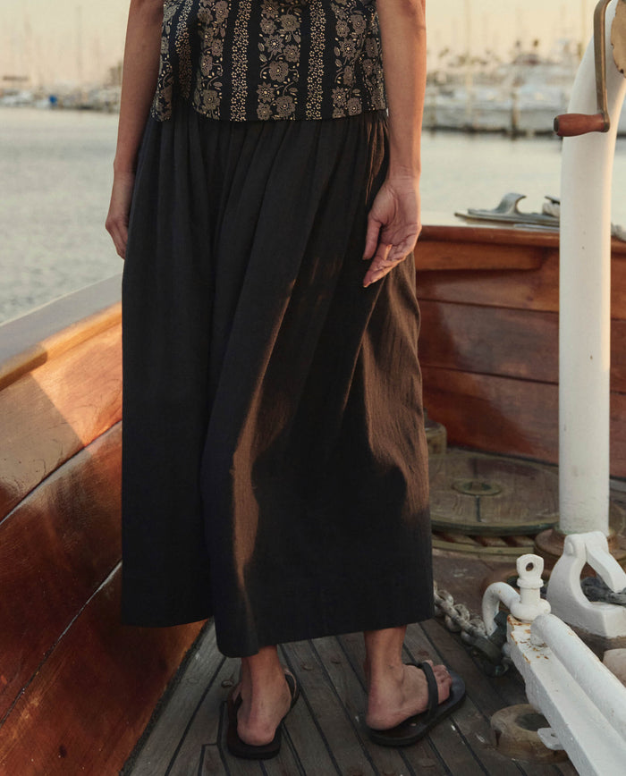 Wearing a patterned top, THE GREAT. SU26 The Viola Skirt in Black, and flip-flops, a person stands on a wooden boat near the water with docked boats visible in the background.