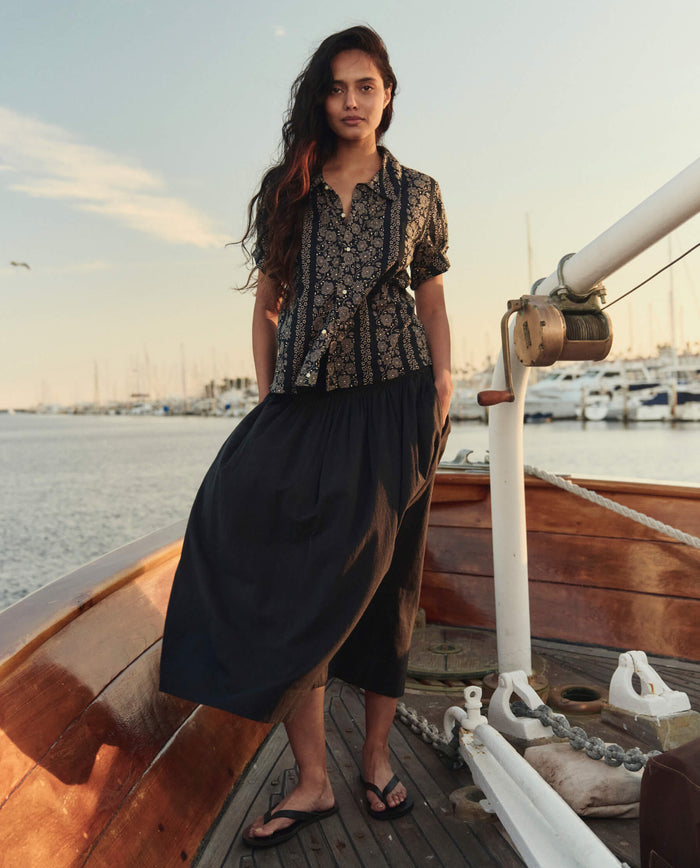 A woman stands on a wooden boat dock at the marina, wearing THE GREAT. SU26’s The Viola Skirt in black, a patterned short-sleeve shirt, and flip-flops. Sailboats and water create a scenic backdrop.