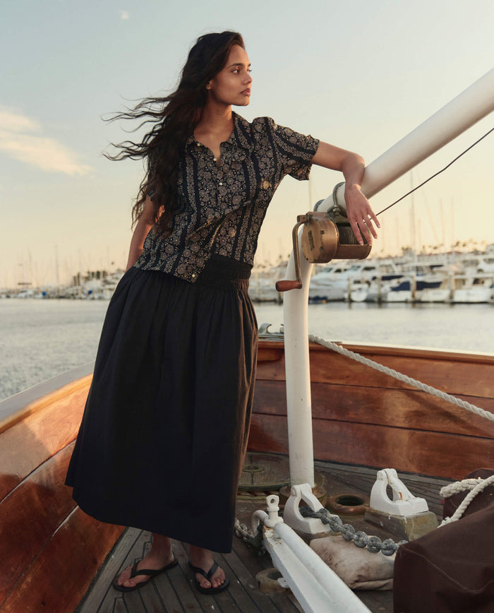 A woman stands on a wooden boat in front of a marina, wearing THE GREAT. SU26 The Viola Skirt in Black, paired with a patterned blouse and sandals.