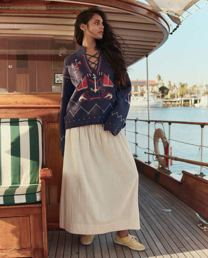 A woman stands on a wooden boat deck wearing a navy sweater with red and white patterns and THE GREAT. SU26 The Viola Skirt in Natural, with water and palm trees in the background.
