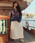 A woman stands on a wooden boat deck wearing a navy sweater with red and white patterns and THE GREAT. SU26 The Viola Skirt in Natural, with water and palm trees in the background.
