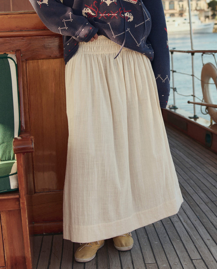 Wearing THE GREAT. SU26's The Viola Skirt in Natural and tan shoes, a person stands on a wooden boat deck with part of a patterned navy sweater showing.