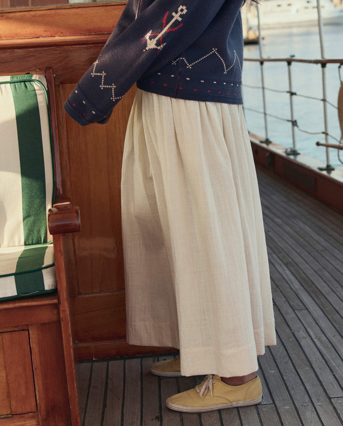 A person stands on a boat deck wearing THE GREAT. SU26’s The Viola Skirt in Natural, a navy sweater with white anchors, and yellow shoes.