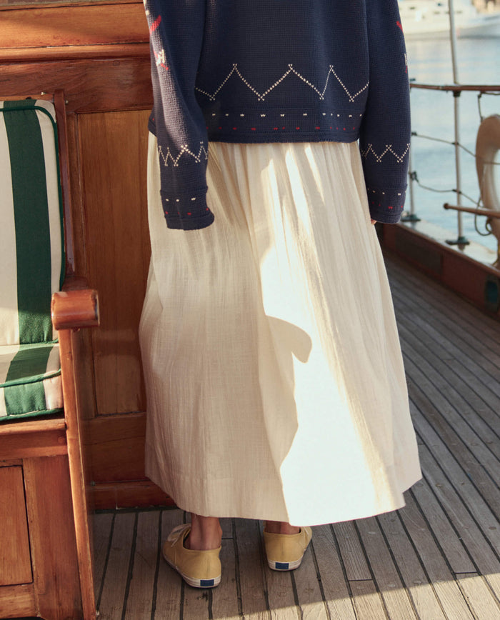 A person stands on a boat deck wearing a navy sweater, beige shoes, and the crisp white The Viola Skirt in Natural by THE GREAT. SU26.