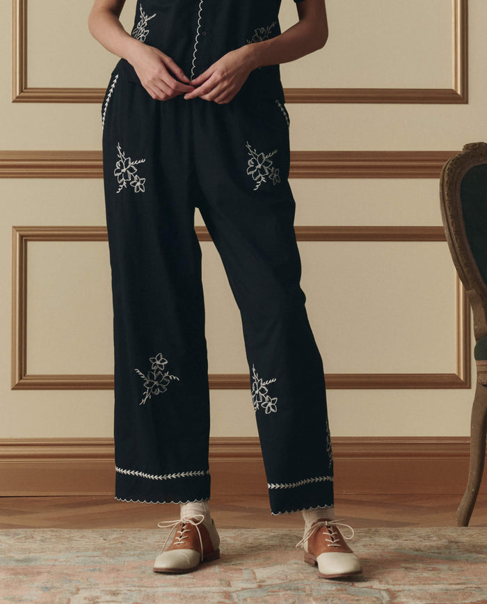 A person stands indoors on a patterned rug near a green upholstered chair, wearing THE GREAT. SP26 D2 The Weekend Pant W/ Hanky Embroidery in black and two-tone brown and white shoes.