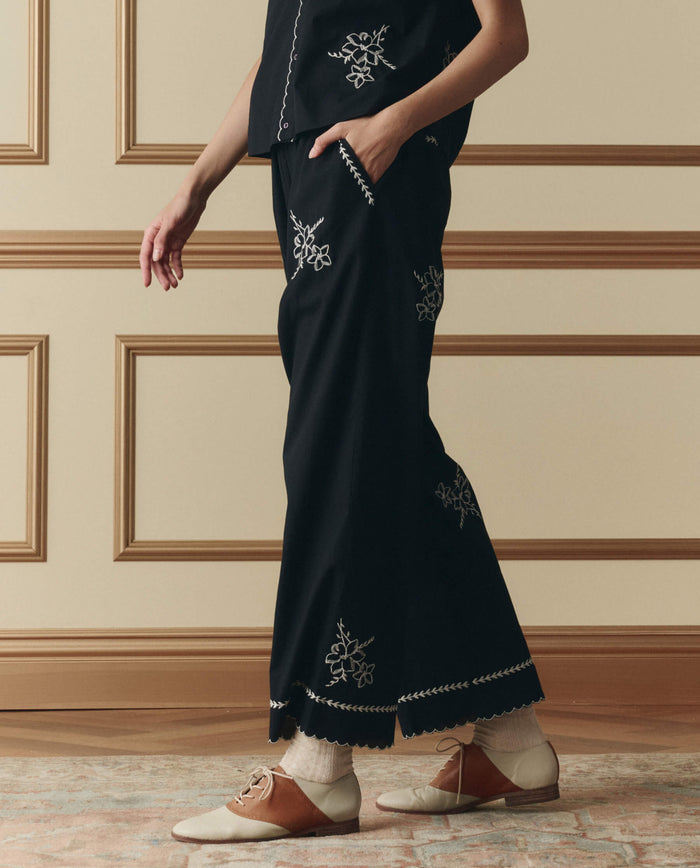 A person in THE GREAT. SP26 D2's The Weekend Pant W/ Hanky Embroidery in black, paired with a matching top, white socks, and tan and white oxford shoes, stands in a room with paneled walls.