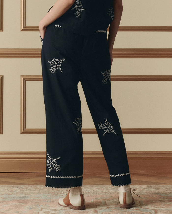 A person stands with their back to the camera, wearing THE GREAT. SP26 D2 The Weekend Pant W/ Hanky Embroidery in black, featuring white floral designs and light-colored shoes, set against a beige wall with decorative molding.