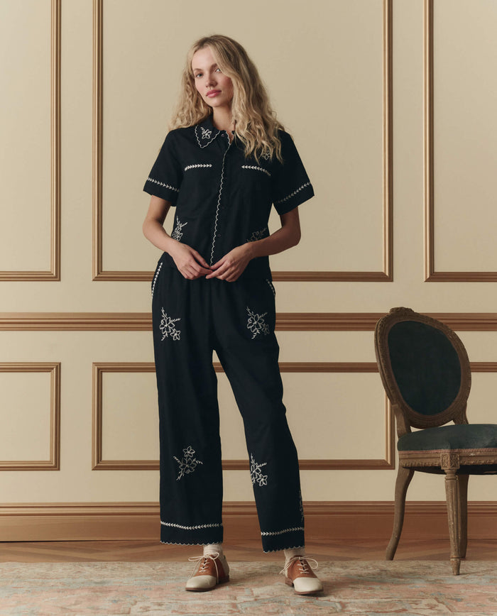 A woman stands indoors in front of a beige paneled wall next to a vintage chair, wearing THE GREAT. SP26 D2 The Weekend Pant W/ Hanky Embroidery in black with white detailing and brown and white shoes.