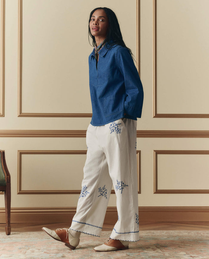 A woman stands indoors, facing forward in a relaxed pose, wearing THE GREAT. SP26 D2 The Weekend Pant w/ Hanky Embroidery in Cream and beige flats, paired with a blue long-sleeve shirt.
