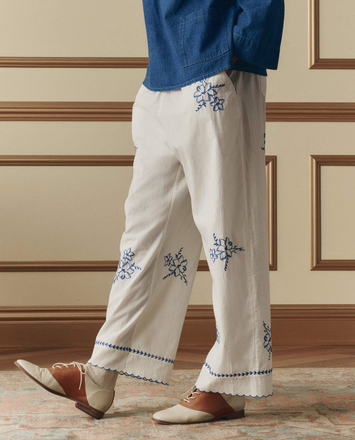 A person wears THE GREAT. SP26 D2 The Weekend Pant W/ Hanky Embroidery in cream, paired with a blue top, beige socks, and brown-and-white shoes, standing on a patterned rug by a paneled wall.
