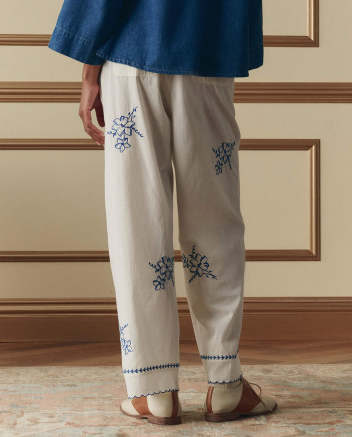 A person stands indoors wearing THE GREAT. SP26 D2's The Weekend Pant W/ Hanky Embroidery in cream, paired with a blue top and two-tone shoes. The photo from behind highlights the intricate blue floral hanky embroidery.