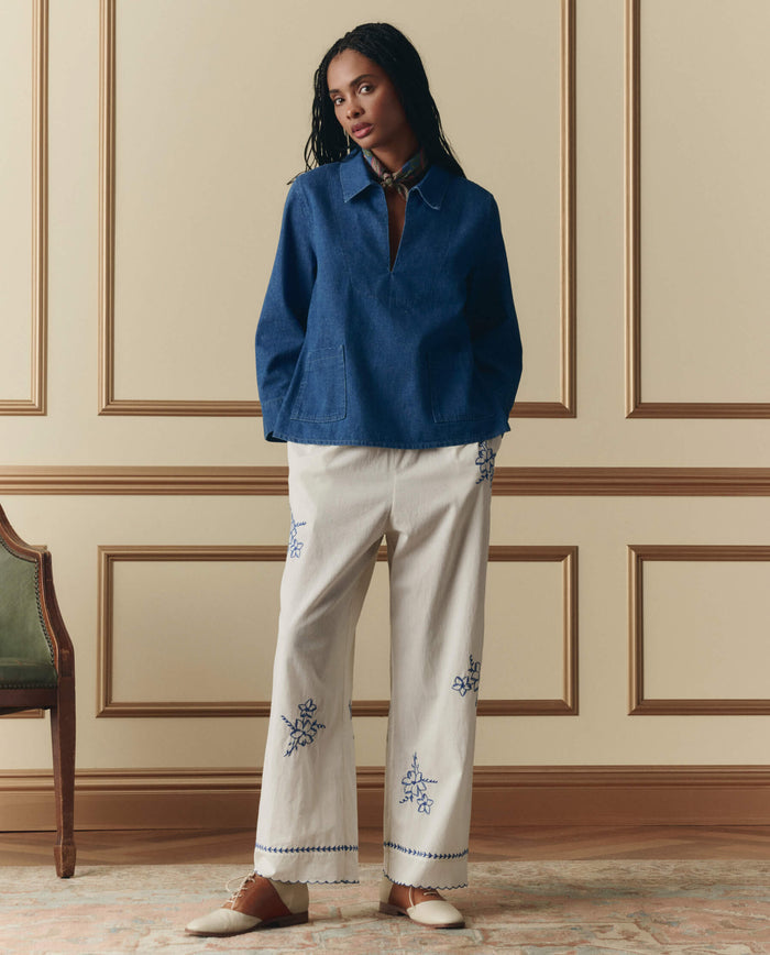 Indoors, a person wears THE GREAT. SP26 D2 The Weekend Pant W/ Hanky Embroidery in cream, styled with a blue long-sleeve top, neck scarf, and white shoes.