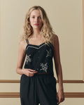A woman with wavy blonde hair wears THE GREAT.'s The Weekend Tank W/ Hanky Embroidery in black, paired with matching pants, standing against a beige wall with decorative molding.