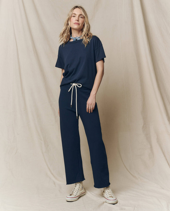 A woman in a blue outfit featuring THE GREAT. FALL 23 KNITS' The Wide Leg Cropped Sweatpant in True Navy and a 100% cotton T-shirt stands on a cream backdrop, complemented by her blue neck scarf and white sneakers.