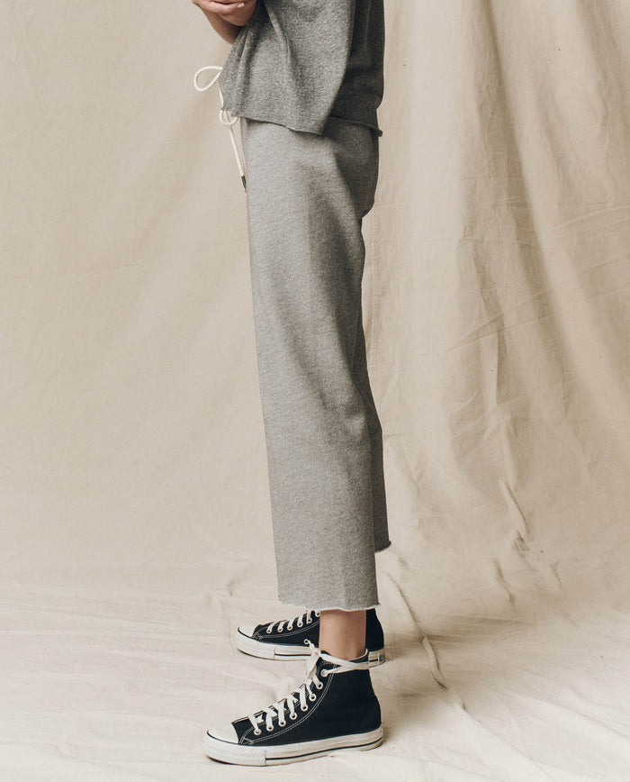A person stands on a draped fabric backdrop, dressed in The Wide Leg Cropped Sweatpant - Varsity Grey from THE GREAT. CORE KNITS and black high-top sneakers with white soles.