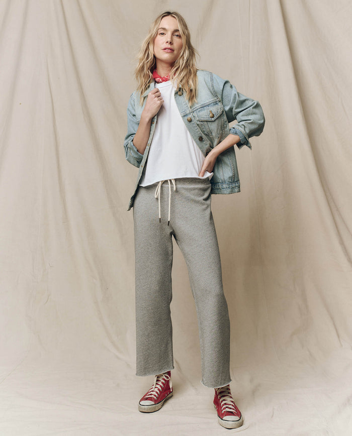 A person poses against a beige fabric backdrop wearing THE GREAT. CORE KNITS The Wide Leg Cropped Sweatpant in Varsity Grey, a denim jacket, white t-shirt, and red sneakers.