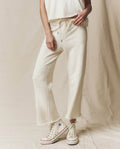 Someone wearing The Wide Leg Cropped Sweatpant in Washed White by THE GREAT. CORE KNITS and a sleeveless top, paired with white high-top sneakers, stands on a neutral fabric backdrop.