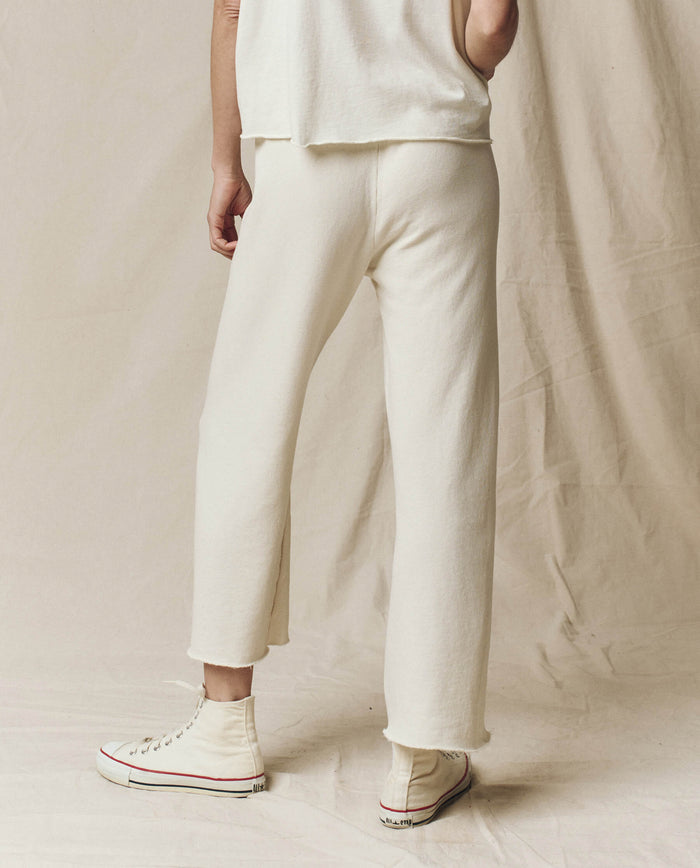A person in washed white wide leg cropped sweatpants by THE GREAT. CORE KNITS stands on a canvas backdrop, facing away. Their outfit is made of 100% cotton for comfort and is paired with white high-top sneakers featuring red and black details.