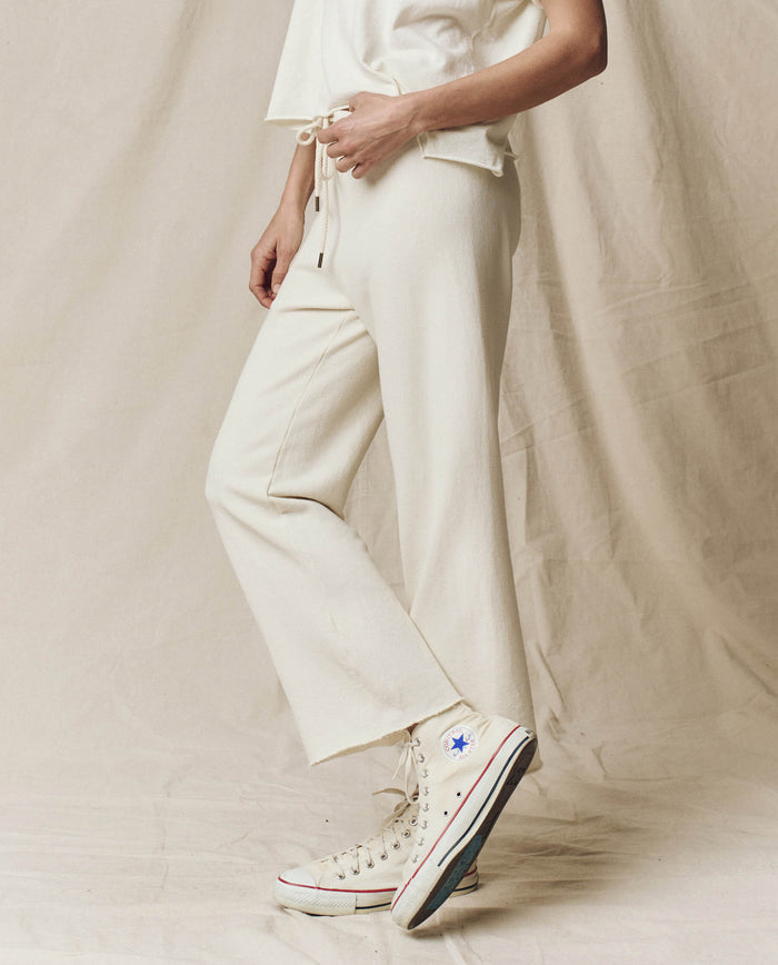 A person in "The Wide Leg Cropped Sweatpant" by THE GREAT. CORE KNITS, crafted from 100% cotton for comfort, stands against a beige background wearing the washed white style with a drawstring waist and wide cropped legs, paired with sneakers.