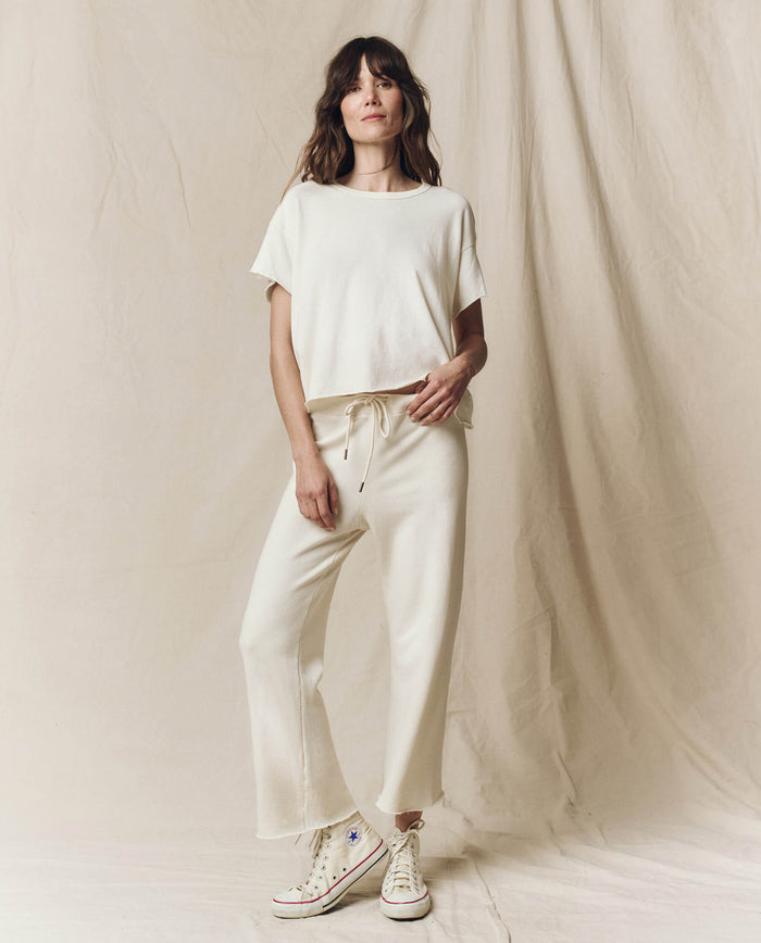 Someone in The Wide Leg Cropped Sweatpant by THE GREAT. CORE KNITS, in Washed White, paired with sneakers stands against a beige fabric backdrop.