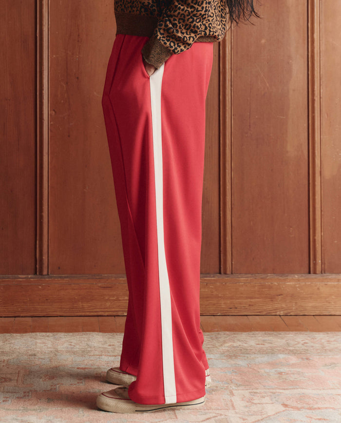 A person wears THE GREAT. SP26 D1 The Wide Leg Track Pant in Poppy Red with a white side stripe, beige shoes, and a leopard print top, standing indoors on a patterned rug against a wooden wall background.