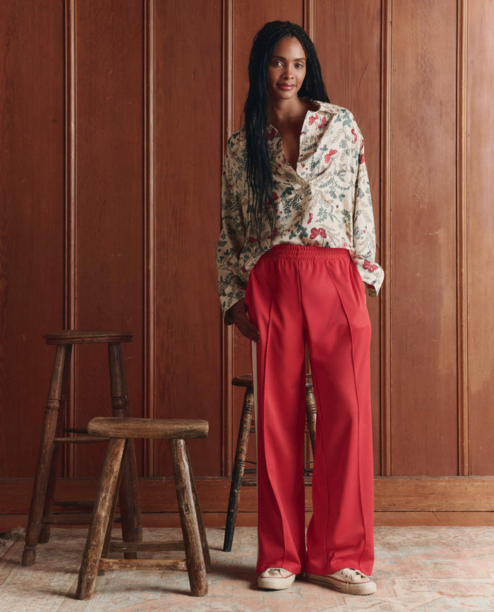 Wearing THE GREAT. SP26 D1 The Wide Leg Track Pant in Poppy Red, a person with long braids and a floral blouse stands indoors by wooden stools against a wood-paneled wall, paired with white shoes.