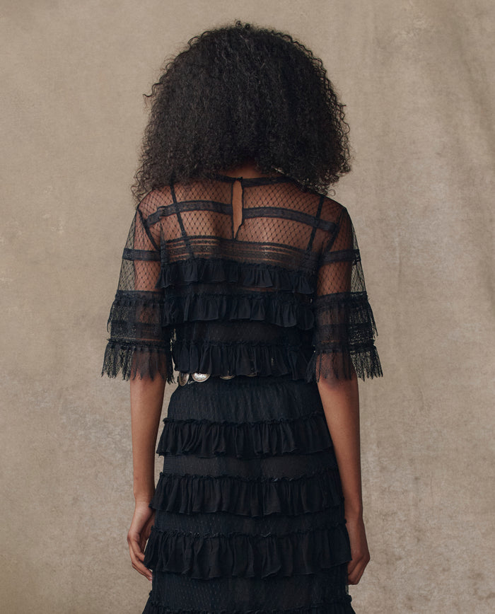 A person with curly hair faces away, wearing THE GREAT. H25 COLLECTION's The Winter Mesh Flounce Top in black—a sheer, ruffled, short-sleeved piece—set against a neutral background.