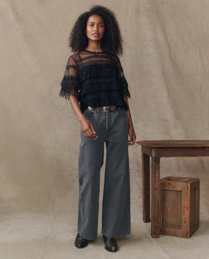 A woman stands indoors in THE GREAT. H25 COLLECTION’s The Winter Mesh Flounce Top in black, styled with high-waisted wide-leg jeans, black shoes, and a belt beside a wooden table and crate—a chic winter look.