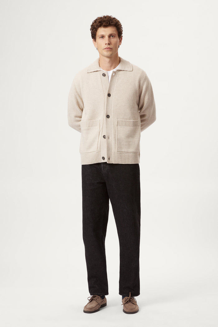 Man wearing a beige woolen cardigan, black pants, and brown shoes on a white background