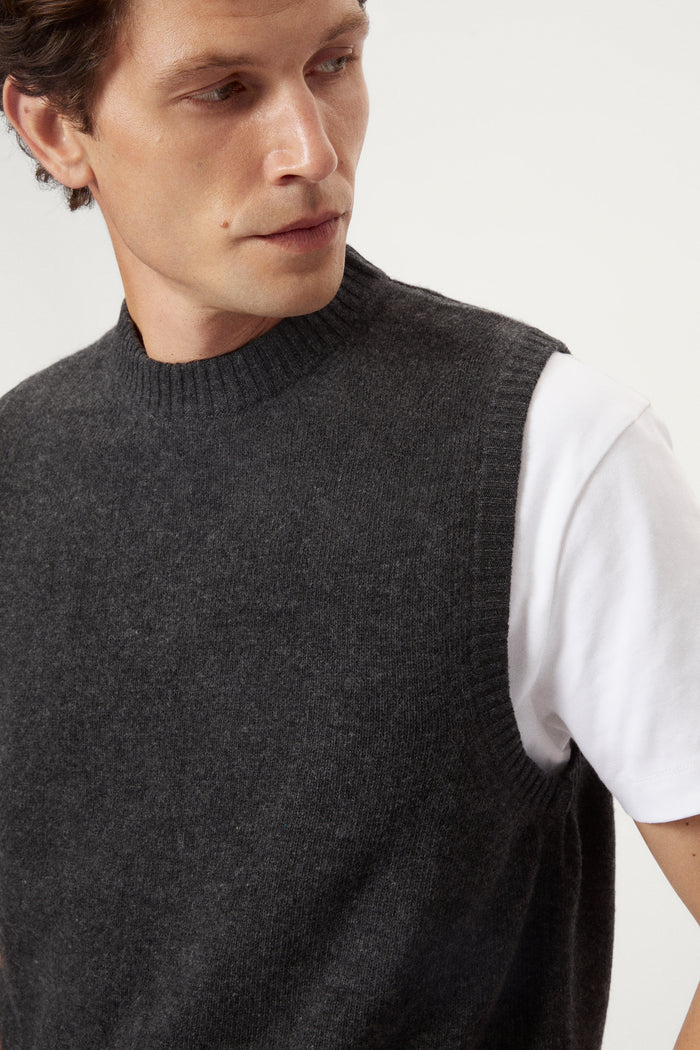 Man wearing a dark gray knitted vest over a white shirt on a light background