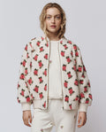 A woman wears THE GREAT. H25 ROSEBUD “The Zip Front Plush Bomber – Cream Rose” with matching pants and a white tee, all featuring pink rose prints and an embroidered motif, standing against a plain background.
