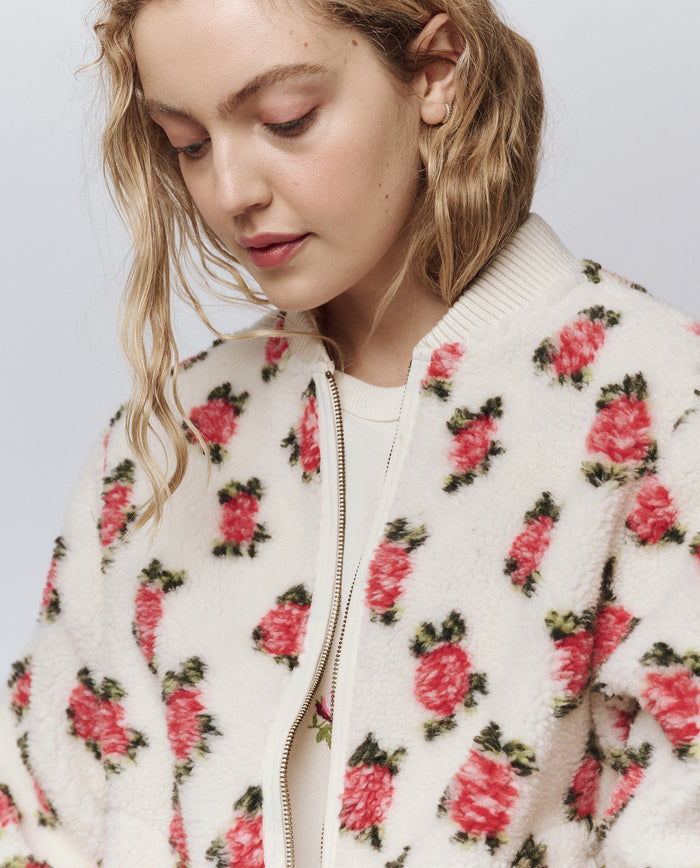 A person with wavy blonde hair looks down while wearing THE GREAT. H25 ROSEBUD The Zip Front Plush Bomber -- Cream Rose, featuring a white base with a pink and green floral pattern.