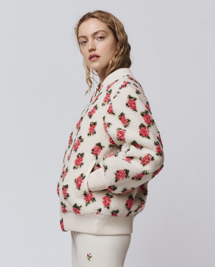 A woman in profile wears THE GREAT. H25 ROSEBUD's The Zip Front Plush Bomber—Cream Rose, featuring an embroidered rose motif, with hands in pockets and paired with cream pants against a plain background.