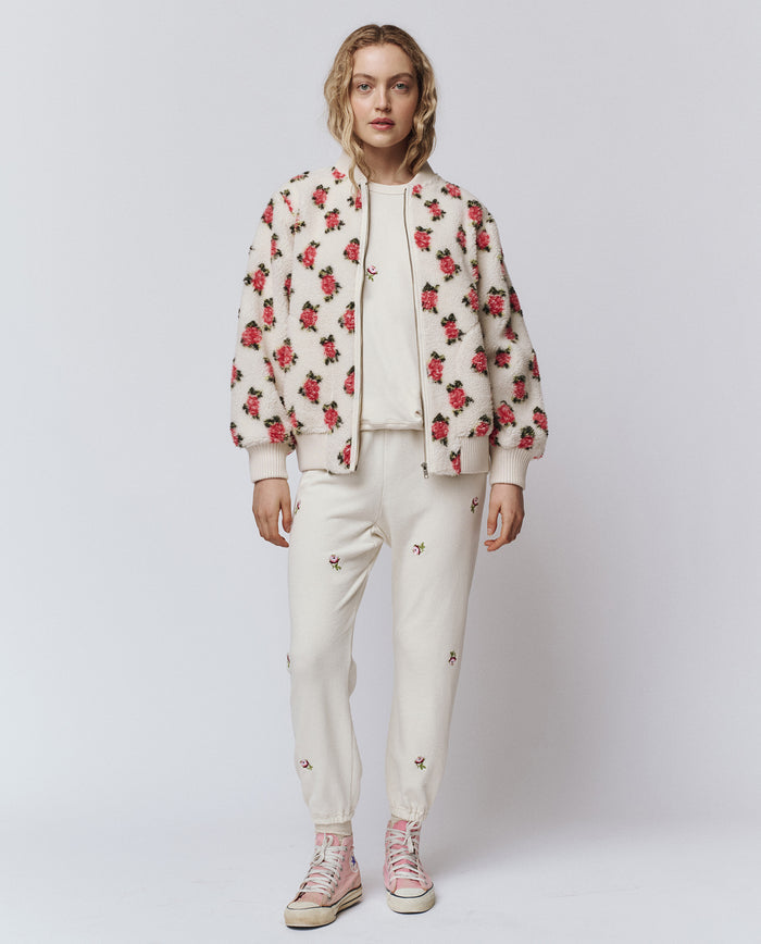 A person stands against a white background wearing THE GREAT. H25 ROSEBUD’s Zip Front Plush Bomber in Cream Rose, featuring an embroidered rose motif, paired with joggers and light pink high-top sneakers.